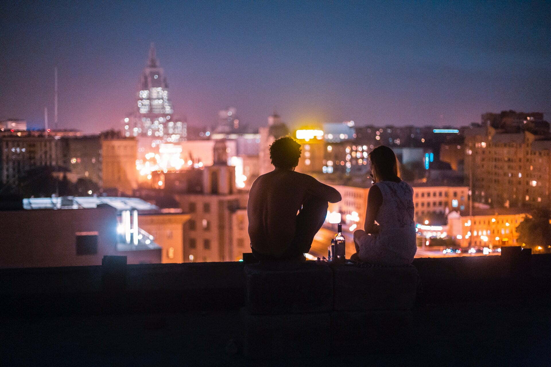 a couple sitting on a ledge overlooking a city at night